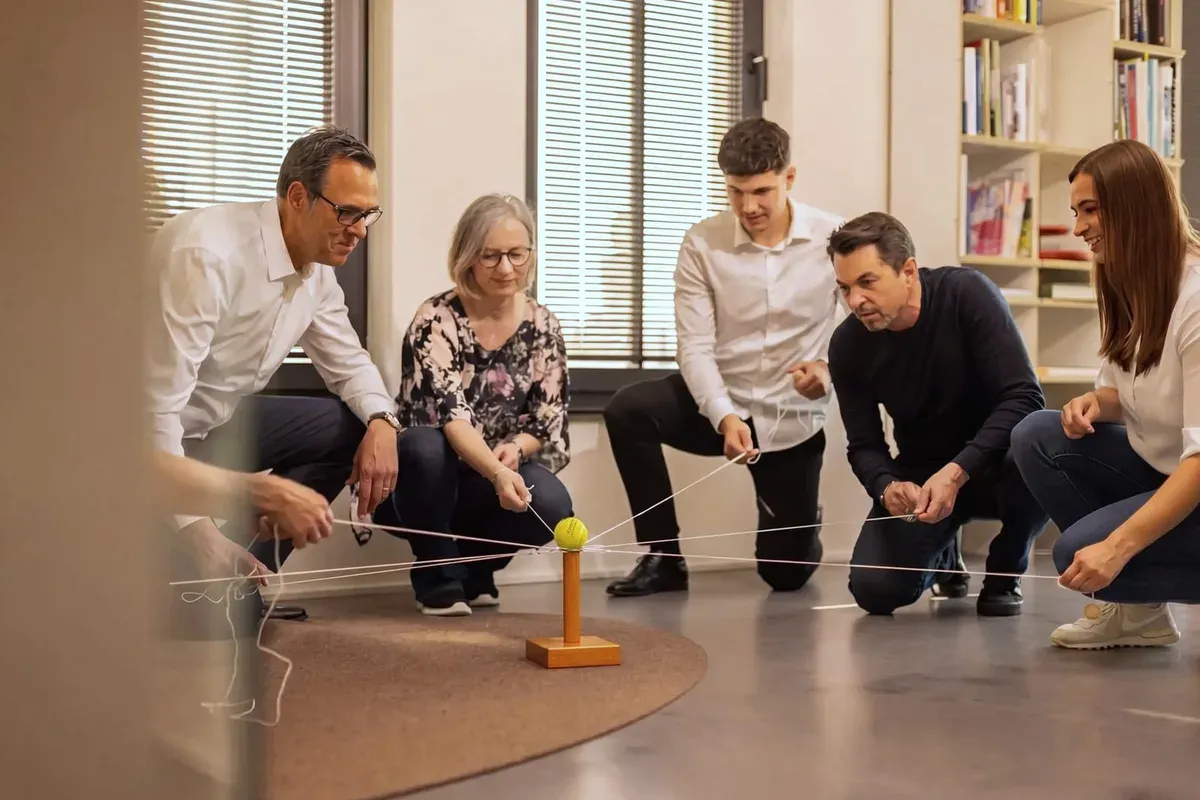 Fünf Führungskräfte balancieren gemeinsam einen Ball auf gespannten Schnüren als Teamübung.